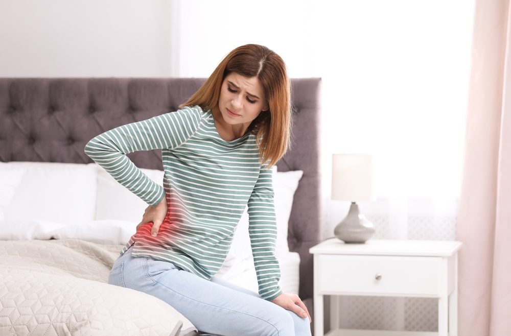 A woman sitting on a bed grimaces in pain and rubs her back while wondering how long after an accident she has to file a claim. 